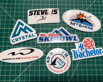 Könnte beinhalten: Eine Sammlung von neun bunten Skigebietsaufklebern. Die Aufkleber zeigen die Namen verschiedener Skigebiete, darunter Stevens Pass, Crystal, Mt. Hood Ski Bowl, Mt. Baker, Whistler Blackcomb, 49° North Mountain Resort und Mt. Bachelor.