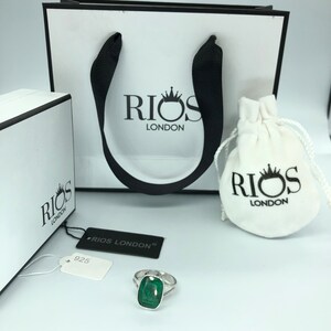 May include: A silver ring with a large, emerald green gemstone. The ring is displayed on a white surface with a white box and a white bag with the logo "RIOS LONDON" printed on it.