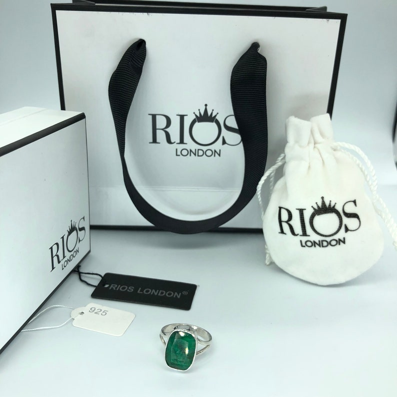 May include: A silver ring with a large, emerald green gemstone. The ring is displayed on a white surface with a white box and a white bag with the logo "RIOS LONDON" printed on it.