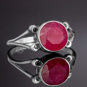 以下が含まれることがあります： 大きな丸い赤いルビーの宝石が付いたシルバーリング。リングはシンプルなバンドで、石の周りに装飾的なデザインが施されています。