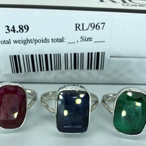 May include: Three silver rings with different coloured gemstones. The rings are on a white surface. The gemstones are red, blue, and green. The rings are for sale for £34.89. The product code is RL/967.