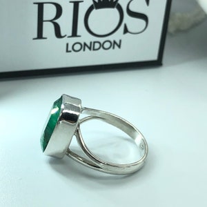 May include: A silver ring with a large, green gemstone. The ring is set with a bezel setting and has a simple band. The ring is made by Rios London.