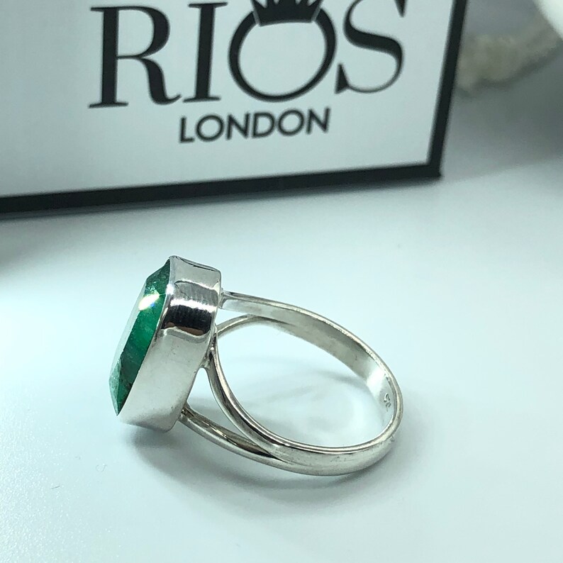 May include: A silver ring with a large, green gemstone. The ring is set with a bezel setting and has a simple band. The ring is made by Rios London.