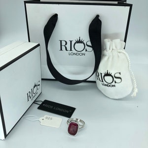 May include: A silver ring with a large, rectangular, red gemstone. The ring is on a white surface with a white tag that says "RIOS LONDON" and "925".
