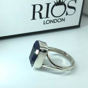 May include: A silver ring with a large, dark blue rectangular gemstone. The ring is set in a simple, modern style. The band is thick and has a slightly rounded shape. The gemstone is set in a bezel setting, which means that the metal surrounds the stone completely. The ring is a size 7.