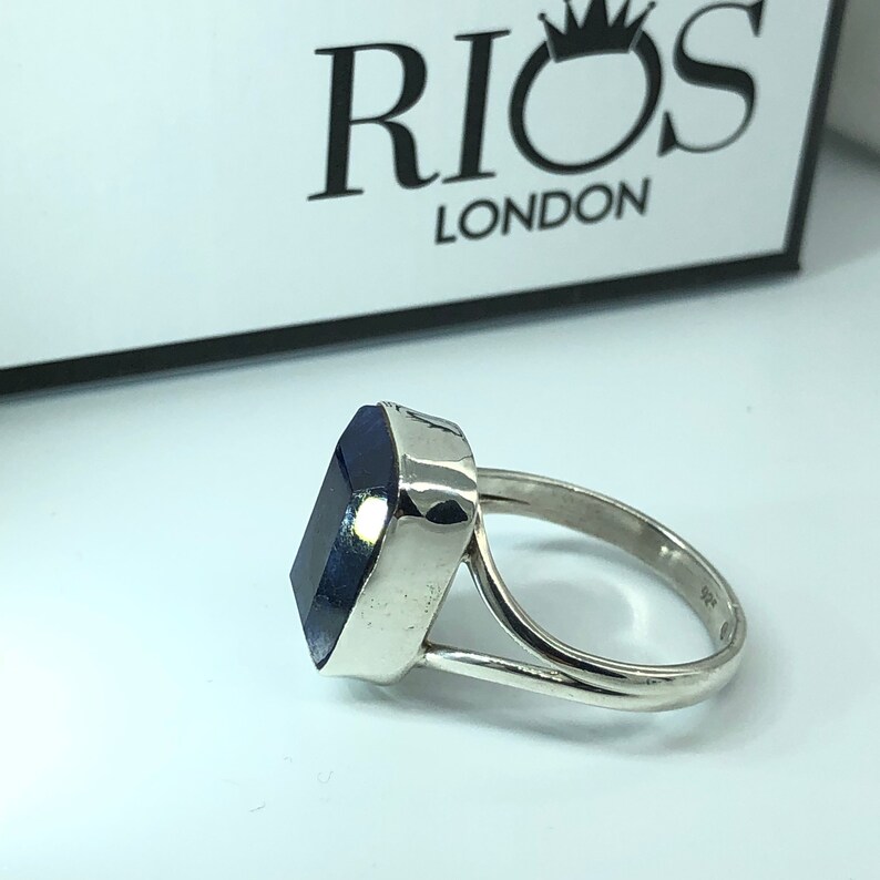 May include: A silver ring with a large, dark blue rectangular gemstone. The ring is set in a simple, modern style. The band is thick and has a slightly rounded shape. The gemstone is set in a bezel setting, which means that the metal surrounds the stone completely. The ring is a size 7.
