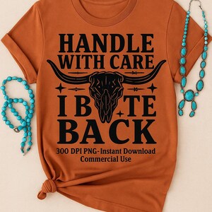 May include: Rust-colored t-shirt with the text "HANDLE WITH CARE IB TE BACK" and a bull skull graphic. Turquoise beaded necklaces are draped on either side of the shirt. The shirt is knotted at the bottom.