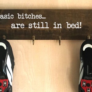 May include: A dark brown wooden sign with the text "basic bitches... are still in bed!" in white lettering. Below the sign are four brass hooks, with two black and white cycling shoes hanging from the hooks.