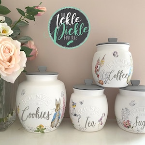 May include: A collection of five white ceramic storage jars with grey lids. Each jar is decorated with floral and bunny illustrations and labelled with the words "Cookies", "Coffee", "Tea", and "Sugar". The jars are from the "Kilner" brand.