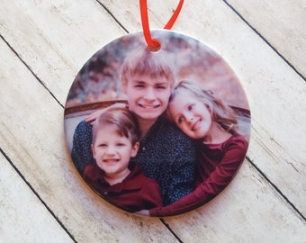 Peut inclure: Une boule de Noël ronde en céramique avec une photo de trois enfants. Les enfants sourient et semblent être des frères et sœurs. La boule est ornée d'un ruban rouge.