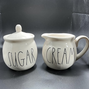 May include: A white ceramic mug with a black handle and the text "But First, Coffee" in a handwritten style. The mug is on a white surface. The mug is approximately 4 inches tall.