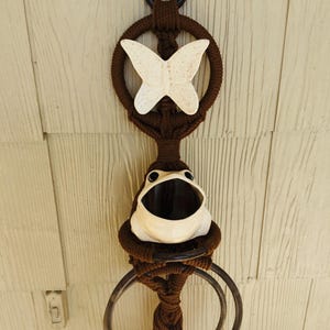 May include: A brown macrame towel rack with a frog figurine and a butterfly decoration. The rack has two metal rings for hanging towels.