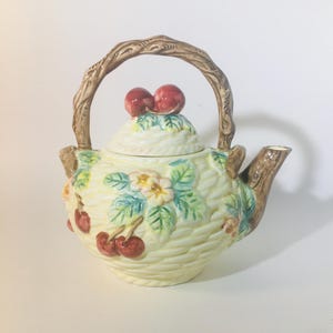 May include: A cream-colored ceramic teapot with a brown branch-like handle and spout. The teapot is decorated with red cherries, green leaves, and small white and yellow flowers. The lid is topped with two red cherries.