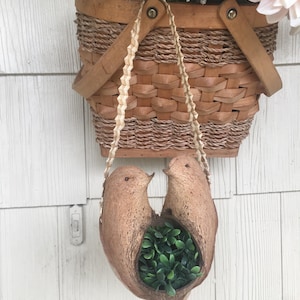 May include: Two coconut shell bird planters hanging from a woven basket with a brown leather handle. The planters are filled with green artificial plants.