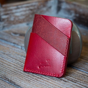 May include: A red leather cardholder with a stitched edge and a small embossed rabbit logo. The cardholder has three angled pockets and is made of different shades of red leather.
