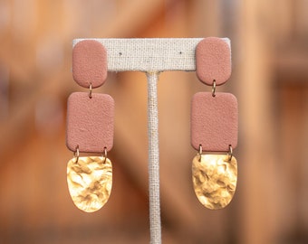 May include: A pair of dangle earrings with a pink clay square and a hammered gold teardrop shape. The earrings are displayed on a white stand.