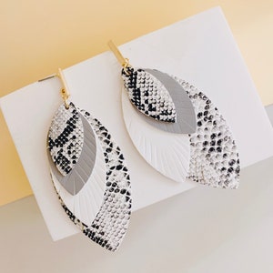 May include: A pair of dangle earrings with a layered design. The earrings are made of white, gray, and black faux leather with a snake skin pattern. The earrings have a gold-tone hook closure.