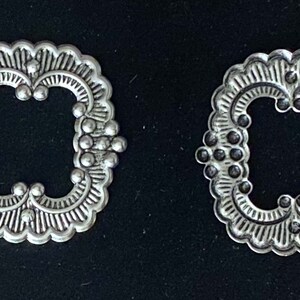 May include: Two silver-colored, ornate buckles with a scalloped edge and a square center opening. Each buckle features a detailed, symmetrical design with small, round embellishments. The buckles are displayed against a dark background.