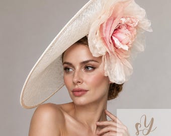 May include: A wide-brimmed cream-colored hat featuring a large, layered, pale pink fabric flower accent on the side. The hat has a textured, woven appearance and is positioned at an angle. The text Sophie Young HATS is visible in the corner.