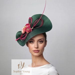 Può includere: Un cappello verde scuro con tesa larga, ornato con nastri rosa e rossi e dettagli floreali. Una lunga e sottile piuma rosa si estende verso l'alto. Un accessorio elegante.