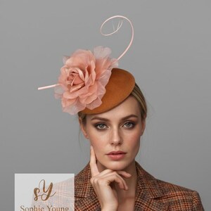 May include: A brown felt hat with a large peach-colored flower and a delicate, curved accent. The hat is adorned with a sheer, layered flower and a thin, curved embellishment. The hat is from Sophie Young Hats.