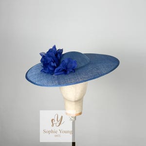 Pu&ograve; includere: Un cappello blu con tesa larga e corona pi&ugrave; piccola, decorato con un grappolo di fiori di stoffa blu. Il cappello &egrave; esposto su una testa di manichino. Il testo "Sophie Young HATS" &egrave; visibile.