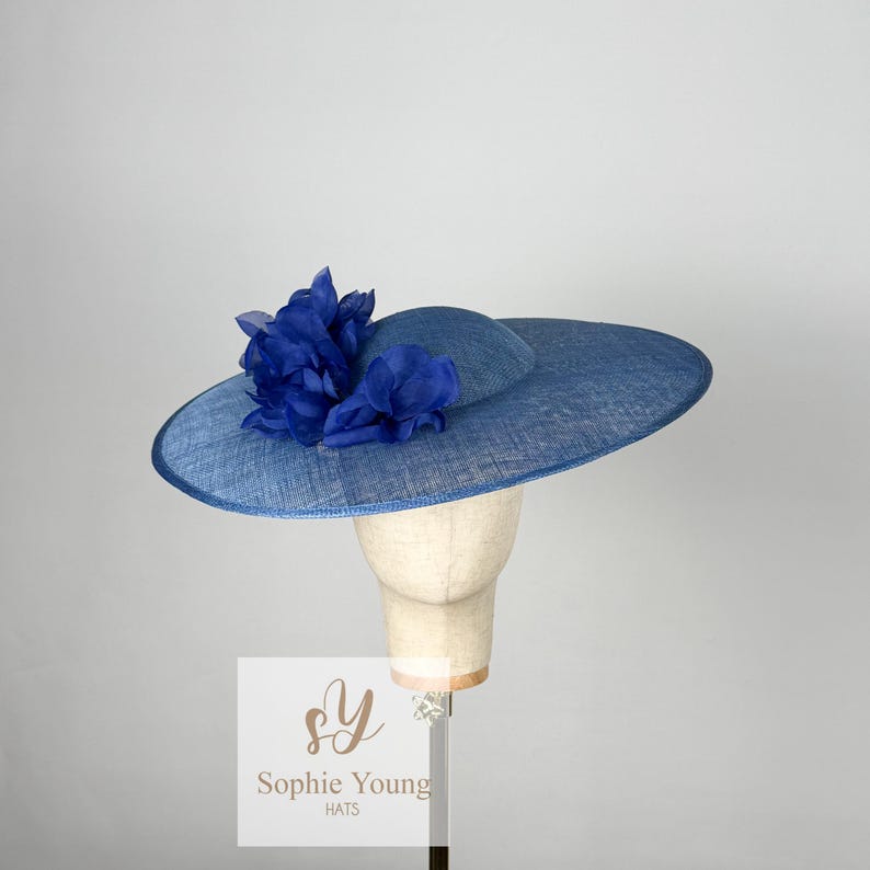 Pu&ograve; includere: Un cappello blu con tesa larga e corona pi&ugrave; piccola, decorato con un grappolo di fiori di stoffa blu. Il cappello &egrave; esposto su una testa di manichino. Il testo "Sophie Young HATS" &egrave; visibile.