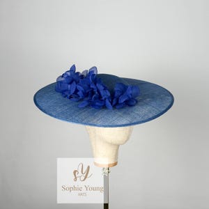 Pu&ograve; includere: Un cappello blu con una tesa larga e un gruppo di fiori di tessuto blu. Il cappello &egrave; esposto su una testa di manichino. Il testo "Sophie Young HATS" &egrave; visibile su un'etichetta bianca.