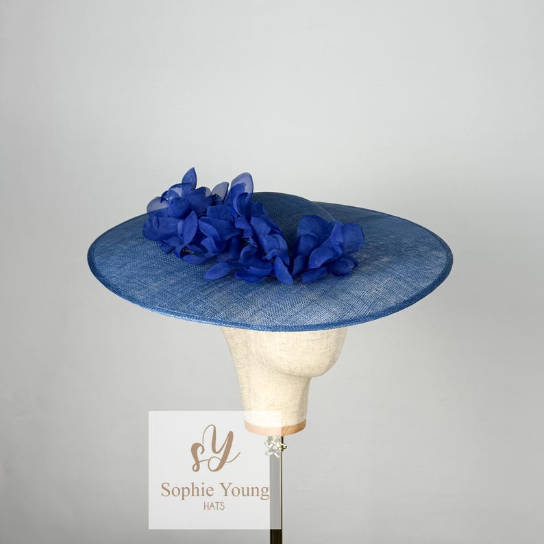 Pu&ograve; includere: Un cappello blu con una tesa larga e un gruppo di fiori di tessuto blu. Il cappello &egrave; esposto su una testa di manichino. Il testo "Sophie Young HATS" &egrave; visibile su un'etichetta bianca.