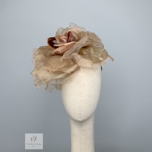 K&ouml;nnte beinhalten: Ein zartes, rosafarbenes Stoffblumen-Haarschmuckst&uuml;ck mit einem mehrschichtigen, gekr&auml;uselten Design. Die Blume besteht aus durchsichtigem Stoff und hat eine leicht strukturierte Oberfl&auml;che.