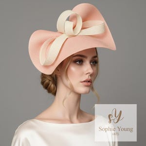 May include: A peach-coloured wide-brimmed hat with a cream-coloured bow and ribbon detail. The hat has a structured design and is from the Sophie Young Hats collection. A stylish accessory for special occasions.