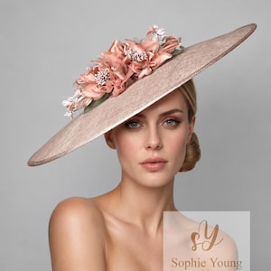 Può includere: Un cappello a tesa larga con una finitura marrone chiaro testurizzata, caratterizzato da un gruppo di fiori in tessuto color pesca e foglie verdi sul lato. Il cappello ha una tesa piatta strutturata e un design pulito. Il logo Sophie Young Hats è visibile.