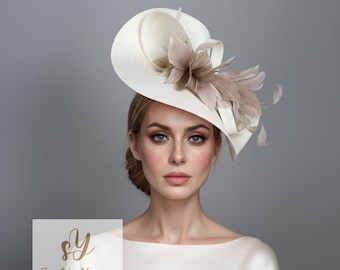 Puede incluir: Un sombrero color crema con ala ancha y un arreglo decorativo de plumas y tela. El sombrero presenta un diseño curvo y está adornado con un grupo de plumas de color marrón claro. El sombrero es de Sophie Young Hats.