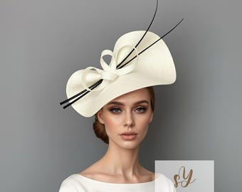 Puede incluir: Un sombrero color crema con ala ancha y lazo decorativo. El sombrero presenta detalles en negro y está diseñado con una forma elegante y asimétrica. Un accesorio sofisticado, perfecto para ocasiones especiales.