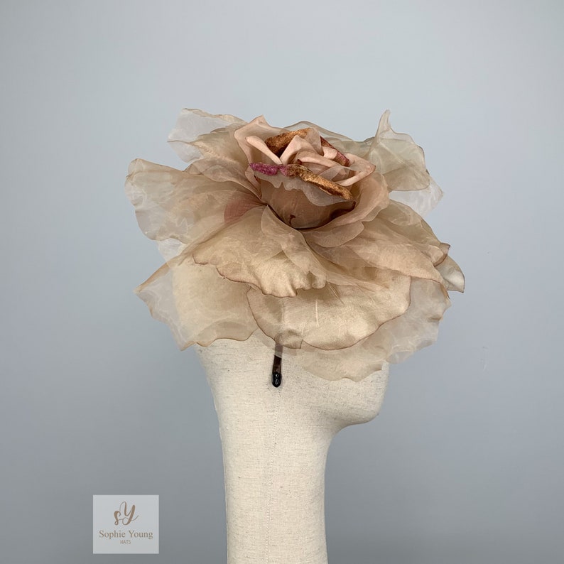 K&ouml;nnte beinhalten: Eine beige Stoffblume mit braunem Zentrum, die als Haarschmuck getragen wird. Die Blume besteht aus mehreren Schichten aus durchsichtigem Stoff und ist an einem schwarzen Haarreif befestigt.