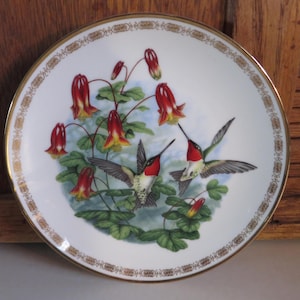 May include: A decorative plate featuring a hummingbird design. The plate has a white background with a gold rim and a detailed illustration of hummingbirds and red and yellow flowers. The hummingbirds are in flight, with their wings spread.