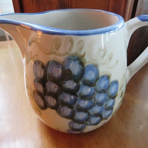 May include: A white ceramic pitcher with a blue rim and a painted design of grapes in shades of blue and green.
