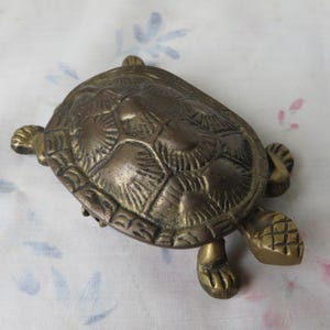 May include: A brass turtle figurine with a detailed, textured shell featuring raised, fan-like patterns on each scute. The metal has a darkened, aged patina, and the turtle is depicted with four legs and a head extending from beneath the shell.