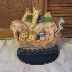 May include: A decorative doorstop shaped like Noah's Ark, featuring pairs of animals including elephants, giraffes, lions, and zebras. The ark is painted in muted tones with a black base and measures approximately 7 inches in length.