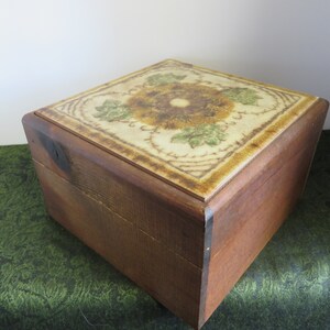 May include: A wooden box with a hinged lid. The lid is decorated with a ceramic tile featuring a floral design in shades of brown, green, and white.