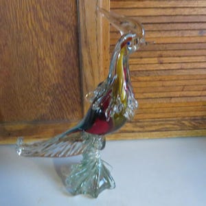 May include: A colorful glass bird figurine with a long neck and tail. The bird is made of clear glass with red, yellow, and blue accents. The bird is standing on a clear glass base.