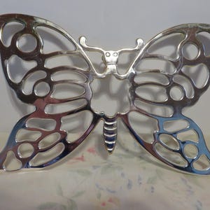May include: A silver-colored butterfly-shaped decorative object. The butterfly has an openwork design with rounded cutouts in the wings and body. The object is made of a shiny, reflective metal and sits on a floral patterned surface.