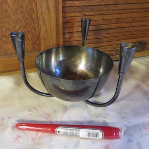 May include: A silver metal candle holder with a central bowl and three candle holders. A red candle is in the foreground. The candle holder has a curved design and is placed on a patterned surface.