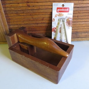 May include: A wooden nutcracker with a hinged lever and a built-in tray for collecting nut shells. The nutcracker is made of dark brown wood and has a natural finish. The lever is made of a lighter wood and has a smooth, rounded handle. The tray is located below the lever and is designed to catch the nut shells as they are cracked. The nutcracker is a traditional kitchen tool that is used to crack nuts, such as walnuts, pecans, and almonds.