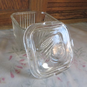 May include: A clear, square glass container with a lid. The lid features an embossed floral design. The container's sides have vertical lines. The lid is slightly ajar, revealing the interior of the container.