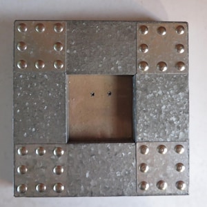 May include: Square metal picture frame with a galvanized finish. The frame is constructed from rectangular sections joined at the corners with metal plates and decorative rivets. The center opening is a square shape.