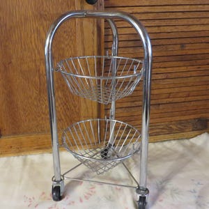 May include: A chrome-plated metal rolling cart with two wire baskets. The cart has a curved handle and sits on four small wheels. The baskets are round and designed to hold items. The cart is suitable for kitchen or bathroom storage.
