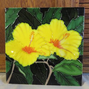 May include: Square ceramic tile featuring two vibrant yellow hibiscus flowers with orange centers, set against a black background with green leaves. The tile has a glossy finish, showcasing the detailed floral design.