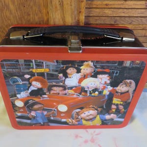 May include: A vintage red metal lunchbox with a black handle and clasp. The front features a colorful illustration of Cabbage Patch Kids in a red car at a drive-in restaurant. The lunchbox has a black top and red trim.
