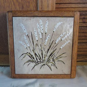 May include: Square wooden trivet with a ceramic tile insert. The tile features a wheat design in white, brown, and green against a beige background. The trivet is framed by a light brown wooden border.